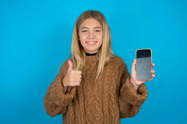 蓝色背景下穿着褐色针织毛衣的金发小女孩展示空白屏幕智能手机 推荐新应用 — 图库照片