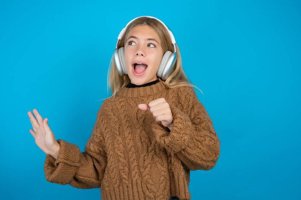 Счастливая блондинка девочка в коричневом трикотажном свитере на синем фоне поет любимую песню держит руку у рта, как будто микрофон носит беспроводные наушники, слушает музыку