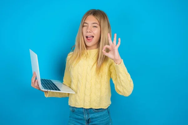 stock image Attractive cheerful skilled beautiful kid girl wearing yellow sweater over blue background using laptop showing ok-sign winkin