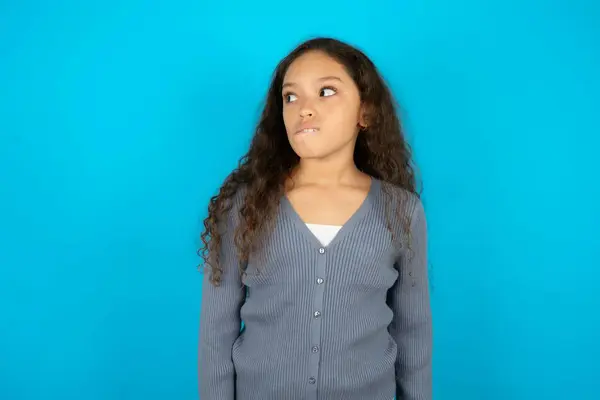 Amazed teenager girl wearing grey sweater against blue background bitting lip and looking tricky to empty space.