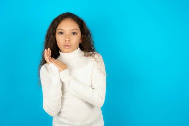 Beyaz kazak giyen duygusal genç kız avuçlarını ovuyor ve kameraya inançsızlıkla bakıyor.