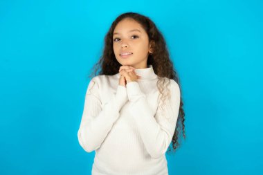 Beyaz kazak giyen mutlu genç kız harika bir şey olacağını umuyor, mutlu bir şekilde yan yana bakıyor, el ele tutuşuyor, mutlu bir yüz ifadesi var..