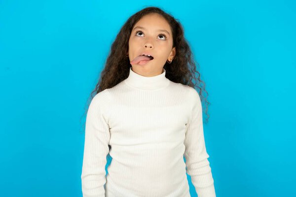 Funny teenager girl wearing white sweater makes grimace and crosses eyes plays fool has fun alone sticks out tongue.