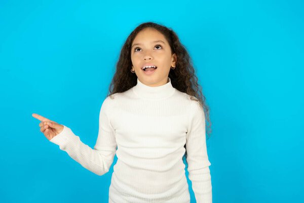 teenager girl wearing white sweater laughs happily points away on blank space demonstrates shopping discount offer, excited by good news or unexpected sale.