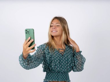 caucasian teen girl wearing green chiffon blouse over white studio background smiling and taking a selfie ready to post it on her social media. clipart