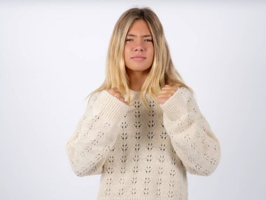 Displeased annoyed caucasian teen girl wearing white knitted sweater over white studio background clenches fists, gestures pissed, ready to revenge, looks with aggression at camera stands full of hate, being pressured clipart