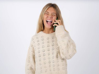 Overemotive happy caucasian teen girl wearing white knitted sweater over white studio background laughs out positively hears funny story from friend during telephone conversation clipart