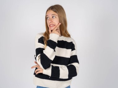 Beyaz stüdyo arka planında çizgili süveter giyen güzel bir genç kız bir soru hakkında endişeli ve gergin bir şekilde düşünüyor..