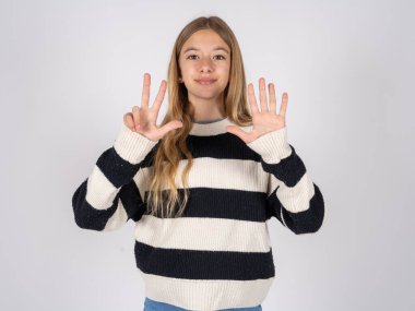 Beyaz stüdyo arka planında çizgili süveter giyen güzel bir genç kız sekiz numaralı parmaklarıyla gülümseyerek kendinden emin ve mutlu bir şekilde onu işaret ediyor..