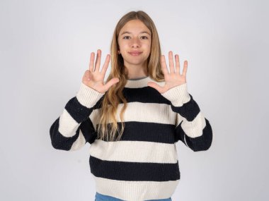 Beyaz stüdyo arka planında çizgili süveter giyen güzel bir genç kız dokuz numaralı parmaklarıyla gülümseyerek kendinden emin ve mutlu olduğunu gösteriyor..