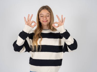 Beyaz stüdyo arka planında çizgili süveter giyen güzel genç kız rahatla ve parmaklarıyla meditasyon hareketi yaparken gözleri kapalı gülümse. Yoga konsepti.