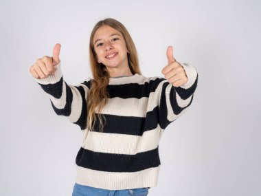 Beyaz stüdyo geçmişine çizgili süveter giyen güzel bir genç kız el hareketleriyle olumlu jest yapmayı onaylıyor, gülümsüyor ve başarı için mutlu. Kazanan jest.