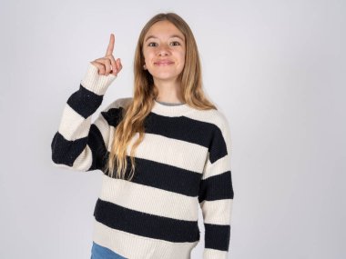 Beyaz stüdyo arka planında çizgili süveter giyen güzel bir genç kız. Parmakla işaret ediyor ve dahiyane düşüncelerden esinlenerek bakıyor. İyi bir fikir işareti gösteriyor.