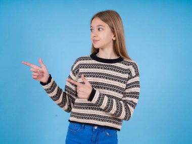 İyimser, beyaz, çizgili süveter giyen güzel bir kız mavi stüdyo arka planında iki eliyle boşluğa bakıyor..