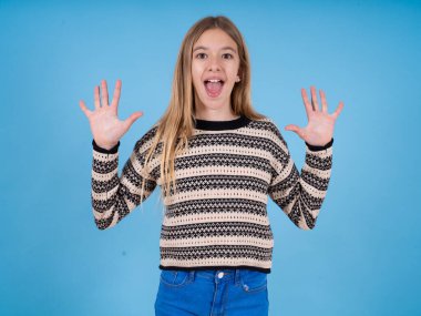 Mavi stüdyo arka planında çizgili süveter giyen güzel beyaz bir kız harika bir sonuç elde ettikten sonra ağzını ve kollarını açar.