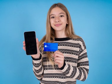 Elinde kredi kartı ve Smartphone ile mavi stüdyo geçmişine çizgili süveter giyen sevimli beyaz bir kızın fotoğrafı. Çevrimiçi alımlar için ayrılmış