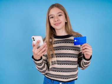 Mavi arka planda çizgili süveter giyen güzel beyaz kız elinde modern cep telefonu ve kredi kartı tutuyor.