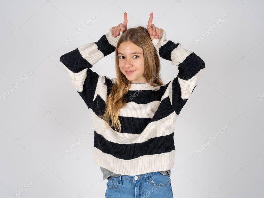 Funny beautiful teen girl wearing striped sweater over white studio background shows horns, fingers on head gesture, posing silly and cute