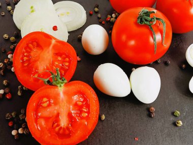 Siyah arkaplanda kırmızı domatesler, soğan halkaları ve soyulmuş bıldırcın yumurtaları ve etrafa saçılmış biber taneleri var..