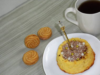 Gri bir masa arkasında çay seremonisi, bir fincan çay, bir kek ve çay tabağında sarı bir kaşık ve yakınlarda yuvarlak kurabiyeler var..