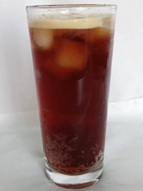 Buzlu koyu bir içecekle dolu uzun bir bardak. İçkinin kırmızımsı kahverengi bir tonu var ve üzerinde birkaç buz küpüyle birlikte açık köpük görülebilir..