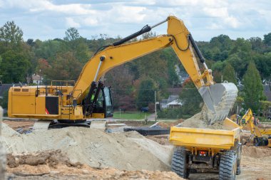 Büyük sarı bir sürüngen kazıcısı ve yanında bir inşaat kamyonu duruyor. Kazıcı çukur kazıyor ve çöp kamyonunu toprak kürekle dolduruyor.
