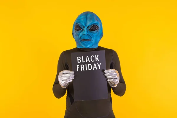 Alien with a blue head holding a black sign with the words \'Black Friday\' in white, on a yellow background. Concept of offers, discounts, opportunities, funny, alien and weird.
