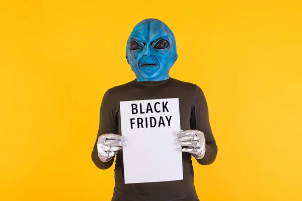 Alien with a blue head holding a white sign with the words \'Black Friday\' in black, on a yellow background. Concept of offers, discounts, opportunities, funny, alien and weird.
