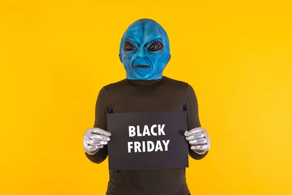 Alien with a blue head holding a black sign with the words \'Black Friday\' in white, on a yellow background. Concept of offers, discounts, opportunities, funny, alien and weird.