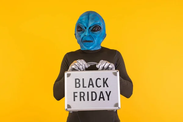 Alien with a blue head holding a metal briefcase with the words \'Black Friday\' on it, on a yellow background. Concept of offers, discounts, opportunities, funny, alien and weird.
