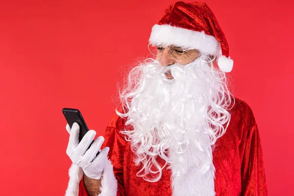 Stock image Man dressed as Santa Claus, consulting his mobile phone, on red background. Christmas, celebration, gifts, consumerism and happiness concept.