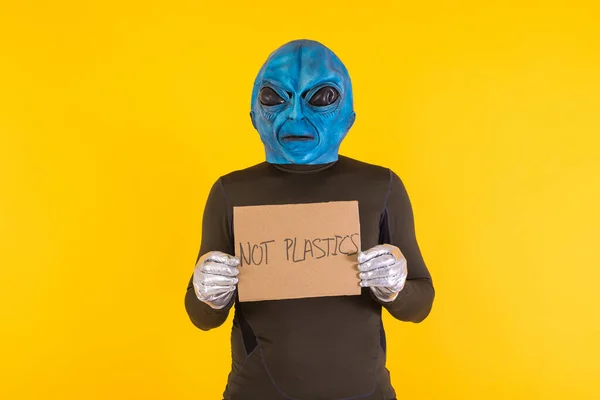 Man with an alien mask with a blue head, holding a cardboard sign that reads: \'No Plastics\'. Concept of bizarre, extraterrestrial, funny, informative, weird and weird.