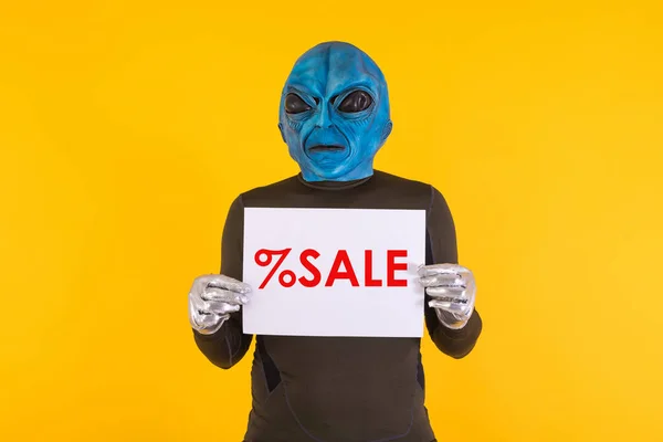 Man with an alien mask with a blue head, holding a white sign with red letters that reads: \'% Sale\'. Concept of bizarre, extraterrestrial, funny, informative, weird and weird.
