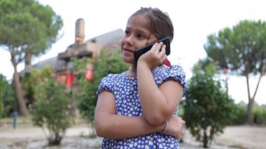 Siyah saçlı, mavi elbiseli, çiçek desenli bir kız sokakta cep telefonuyla konuşuyor. Çocukluk, telefon, teknoloji, internet, mesaj, mutluluk, tatil ve yaşam tarzı kavramı.