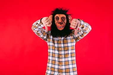 Şempanze maskeli ve kareli gömlekli bir adam, kırmızı arka planda baş parmakları aşağıda..