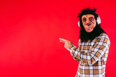 Şempanze maskeli ve kareli gömlekli bir adam, kulaklık takıyor, kırmızı arka planda fotokopi alanını işaret ediyor..