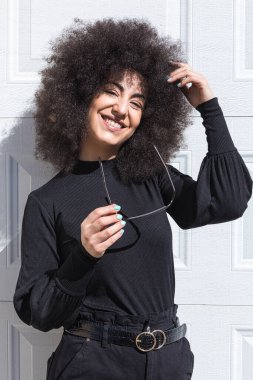 Siyah giyinmiş, beyaz bir kapıya yaslanmış, saçları siyah saçlı genç beyaz bir kadın, gülümsüyor, bandını takıyor, güneşe bakıyor..