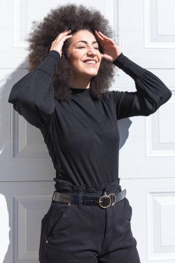 Siyah giyinmiş, beyaz bir kapıya yaslanmış, saçları siyah saçlı genç beyaz bir kadın, gülümsüyor, bandını takıyor, güneşe bakıyor..