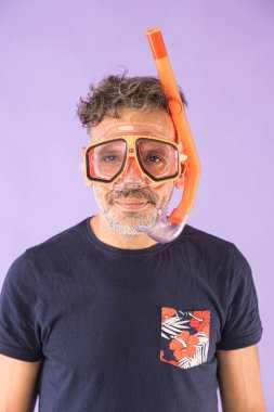 Sakallı, gri saçlı, çiçekli mavi bir tişört giyen ve menekşe arka planda şnorkel gözlüğü ve turuncu bir tüp takan bir adam. Tatil, sahil, macera, eğlence, yaz, turizm.