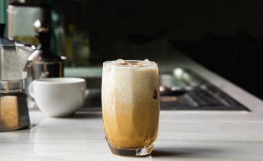 A glass of iced coffee with milk, Asian caffeinated beverages on a marble bar, and a Moka Pot coffee pot in the kitchen.