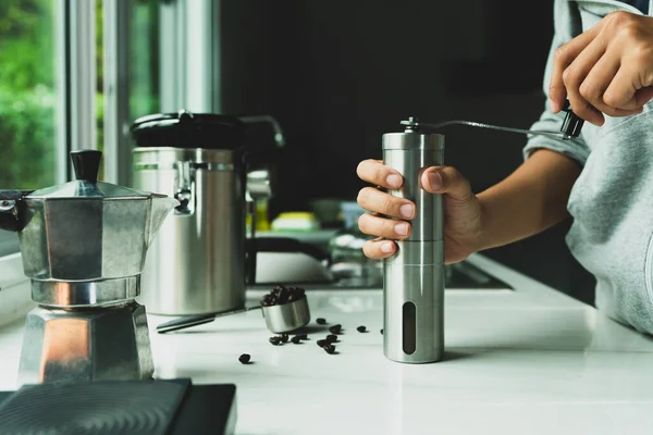 hand is grinding coffee beans with manual stainless steel grinder to make espresso coffee machine, brewing equipment,Vacuum jar collects coffee beans on marble kitchen counter at home in the morning