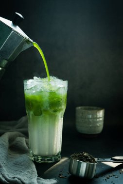 İçinde İtalyan Moka demliği olan Matcha yeşil çayını buzlu süt ve siyah arka planda kaşık üzerine toz yeşil çay ile bir fincana döktü. Metnin için boşluk kopyala