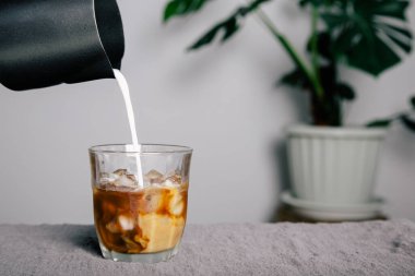 Close-up, black stainless steel pitcher pouring frothed milk over mug into black coffee with ice mug on gray bar with potted plants background.