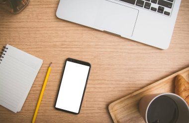 Mobil akıllı telefonun üst görünümü siyah beyaz kullanım için boş kopya alanı oluşturur. Masanın üzerinde dizüstü bilgisayar, kahve kruvasanı, defter ve kalem, ahşap masanın üstünde.