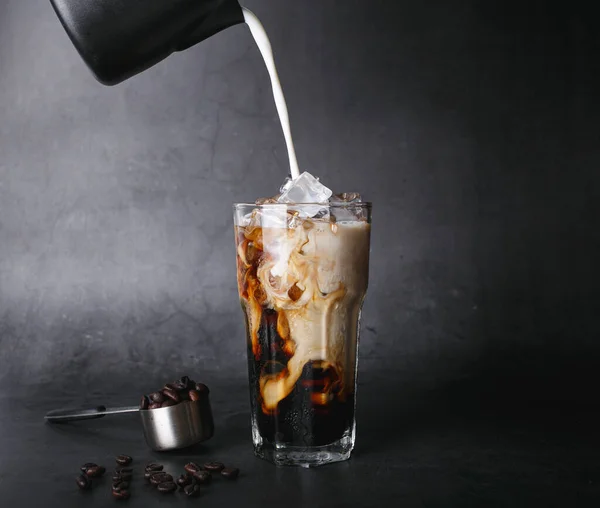 Pouring milk make latte coffee into a transparent glass with ice. and roasted coffee beans on a black background studio photo