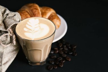 Beyaz fincanda Latte sanat kahvesi, kruvasan, siyah arka planda kavrulmuş fasulye kahvesi, metin alanını kopyala