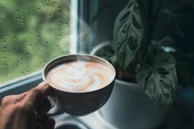 Bir insanın elinde aromatik bir sütlü kahve fincanı var. Pencerenin yanında yağmurlu bir günde kahve iç.