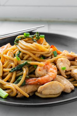 Lezzetli yakisoba eriştesi fesleğenli ve kurutulmuş biberli. İstiridye soslu kızartılmış tavuk fileto, karides, siyah tabakta soğanla süslenmiş. Ev aşçısı dikey fotoğraf 