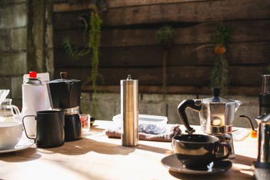Kahve yapma ekipmanları ve aletleri arasında moka demliği, süt köpüğü, kahve öğütücü, bardak, portatif piknik gazı, ahşap masaya yerleştirilmiş.