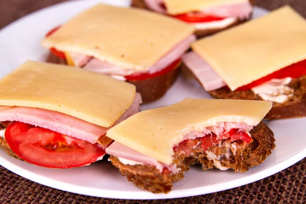 Bitten sandwich with rye bread, tomatoes, ham and cheese on white plate